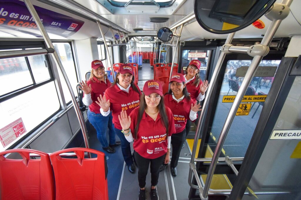 Six women stand inside a bus wearing red shirts and caps with slogans against sexual harassment. They are smiling and raising their hands in a gesture of solidarity. The interior of the bus is modern, with red seats and safety signs visible.