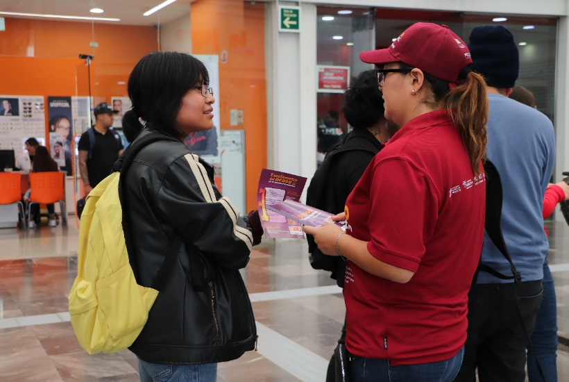A woman wearing a red campaign shirt hands out informational brochures to a passerby inside a public facility.