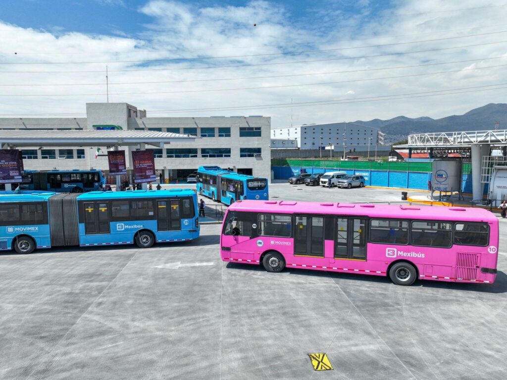 Blue and pink Mexibús units are parked at a transport terminal with modern facilities and mountains visible in the background.