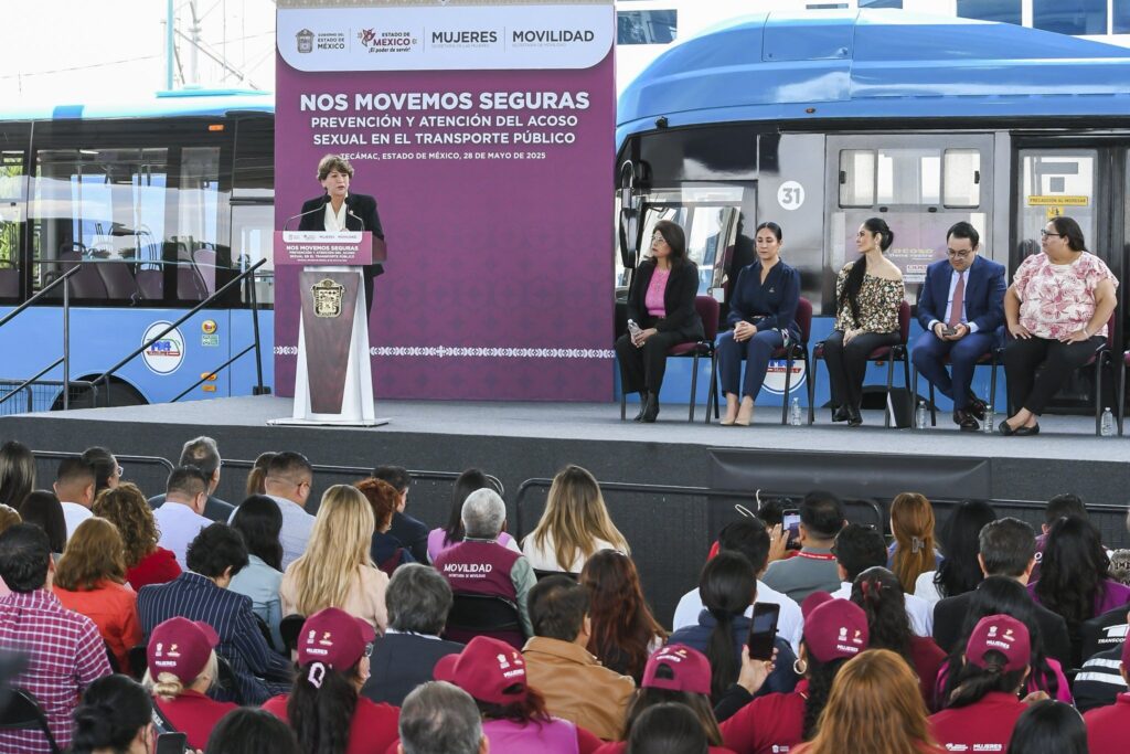 A woman speaks at a podium during a public event with the backdrop reading “Nos Movemos Seguras: Prevención y Atención del Acoso Sexual en el Transporte Público.” Several officials sit behind her on stage, and a blue public bus is parked nearby.