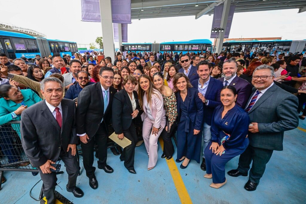 A large group of people poses for a photo at a public transportation event. Several officials stand at the front, smiling at the camera, while a crowd gathers behind them. Blue buses are visible in the background under a covered terminal area.
