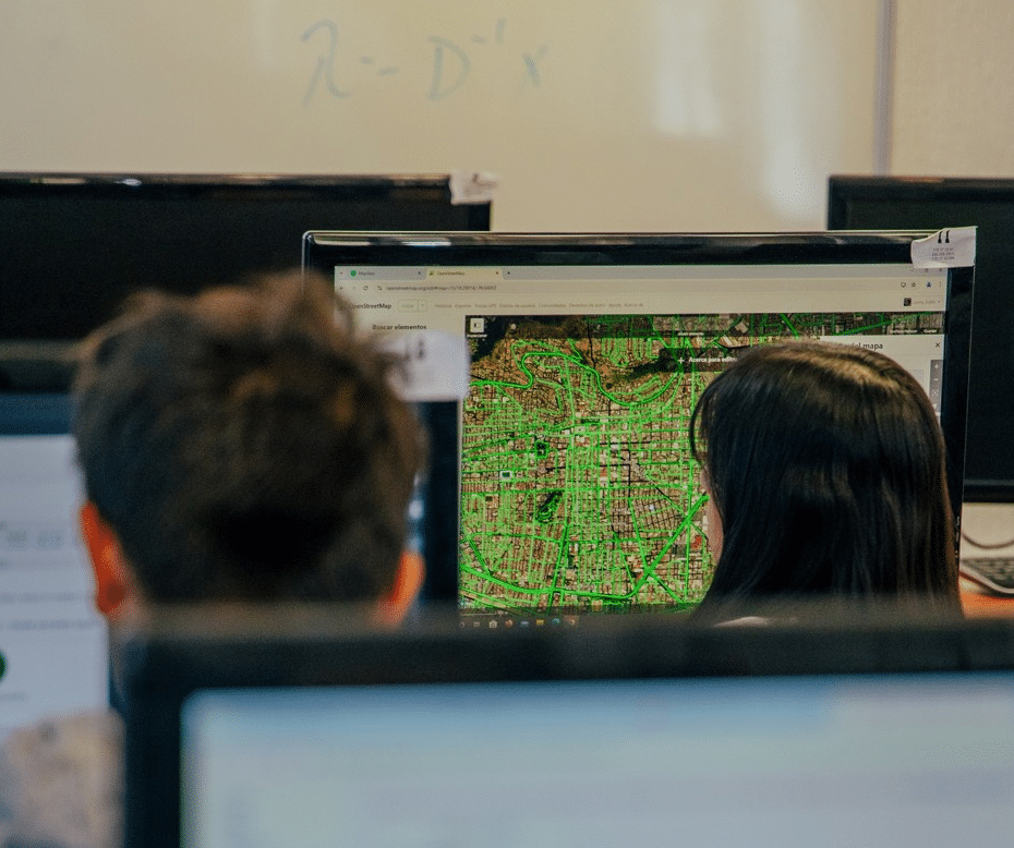 One can see a woman and a man with black hair sitting in front of screens, with an urban map full of green lines on the main screen.