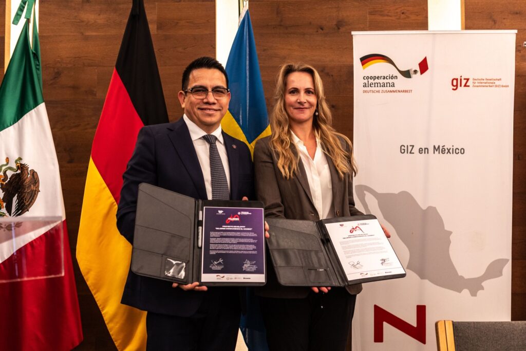 A woman with blond long hair and a man with short dark hair and glasses holding up folders with signed agreement papers in front of the Mexican and German flag and a roll-up with the GIZ cooperation logo and the text "GIZ en México" with a grey Mexico map below.
