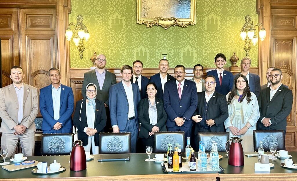 Group picture of 12 men and three women in a historical building with green tapestry including the Hamburg city emblem.