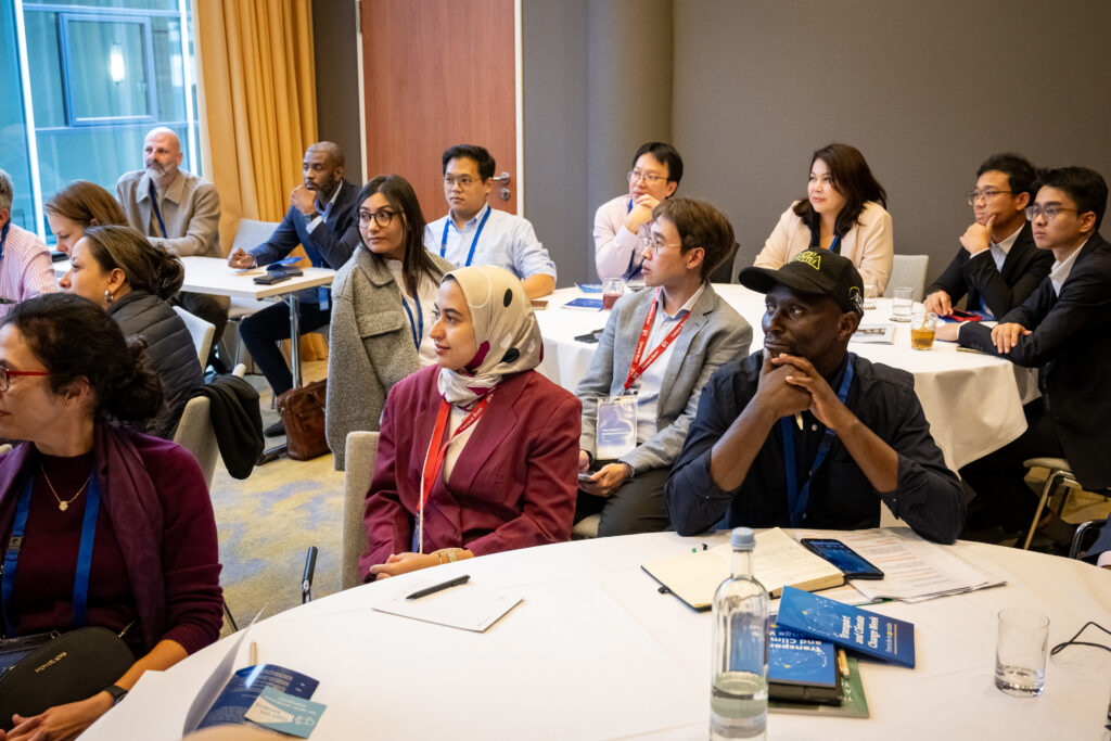 diverse group of conference participants seated at round tables in a meeting room, attentively listening to a presentation. A Transport and Climate Change Week programme booklet is visible on the table. A diverse group of conference participants seated at round tables in a meeting room, attentively listening to a presentation. A Transport and Climate Change Week programme booklet is visible on the table.