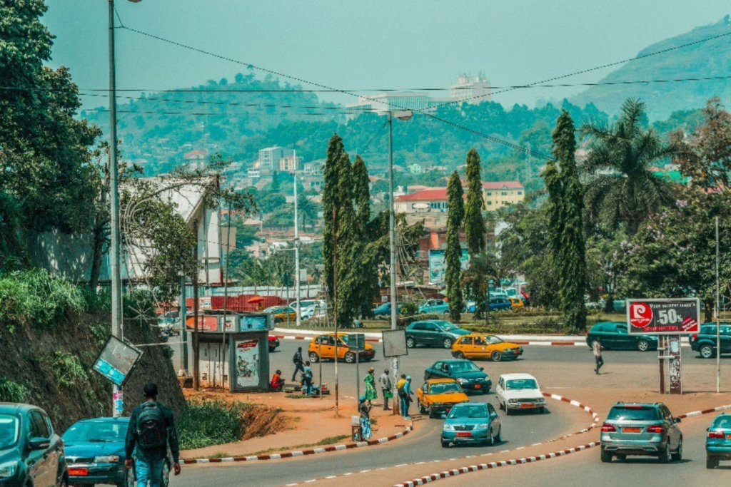 Wide urban road in Yaoundé with cars, taxis and pedestrians, surrounded by green hills and tropical vegetation.