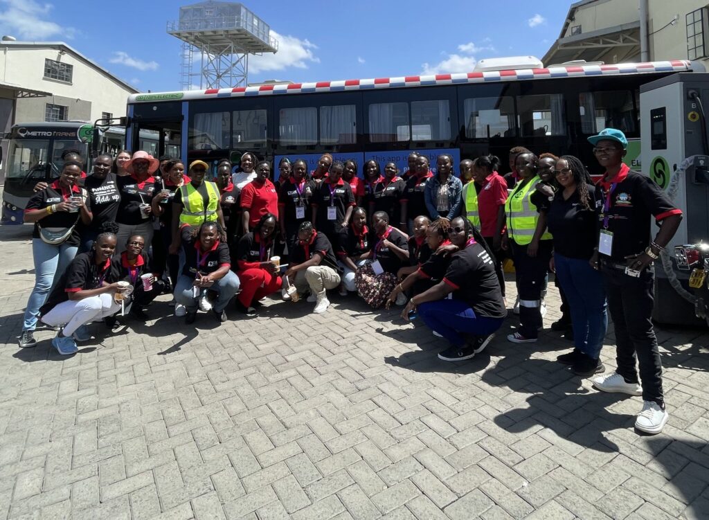A large group of women in matching red and black outfits pose together in front of a blue and white electric bus marked "100% Electric" on a sunny day in Nairobi.